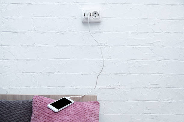 White smartphone lying on the pillow of bed connected to a charging electrical outlet.