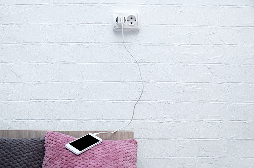 White smartphone lying on the pillow of the bed connected to a charging electrical outlet.