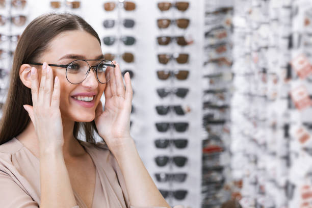Portrait of young lady wearing spectacles in store Ophthalmology And Vision Correction Concept. Portrait of smiling young female client wearing new glasses, choosing frame, standing near rack and showcase with eyewear. Woman trying on spectacles optometrist stock pictures, royalty-free photos & images