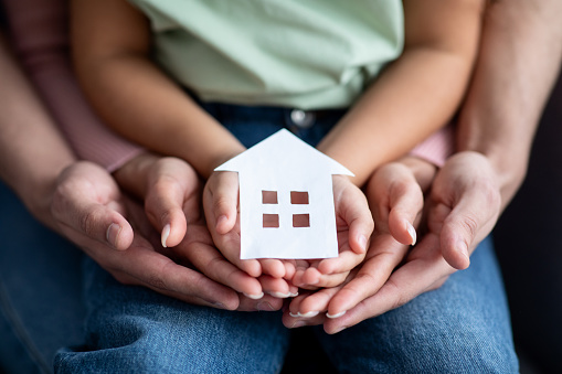 Man, woman and little child holding cutout paper house figure in hands, conceptual image for family housing, home mortgage, real estate, insurance or adoption, closeup shot
