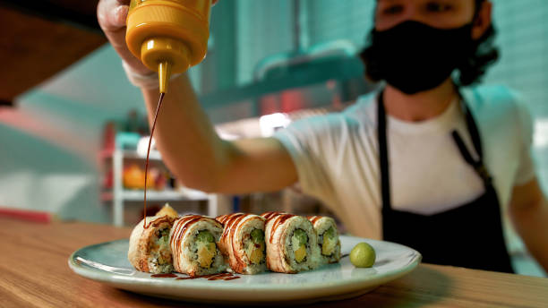 istock Professional sushi chef wearing protective mask adding sauce on top of sushi rolls at commercial kitchen 1305372947