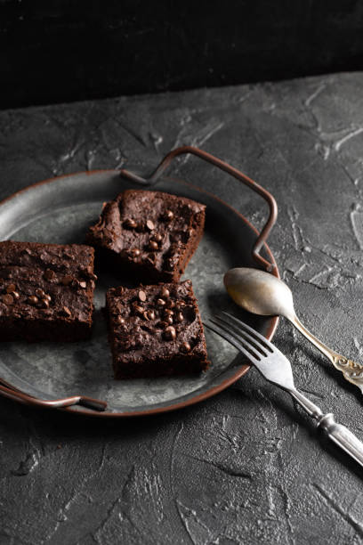 istock Dark cacao brownies with choco drops on old metal tray with vintage silverware on dark background 1305384084