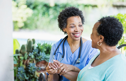 A home healthcare worker, a mature African-American woman in her 50s, visiting a patient on a back yard patio. The patient is a senior African-American woman in her 60s. They are conversing and smiling.