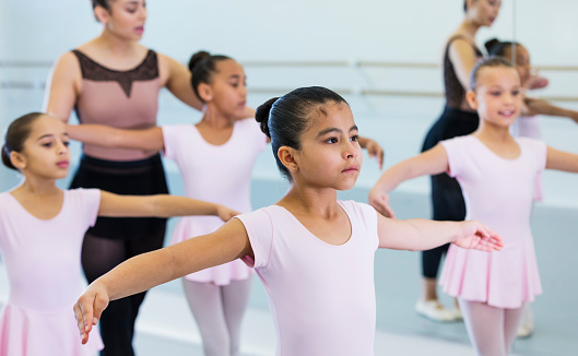 A multi-ethnic group of girls and their dance instructor, in a dance studio, taking a ballet class. The teacher, a young Hispanic woman, is positioning the arms of a 10 year old mixed race African-American and Caucasian girl. The focus is on the 8 year old Hispanic girl in the foreground.