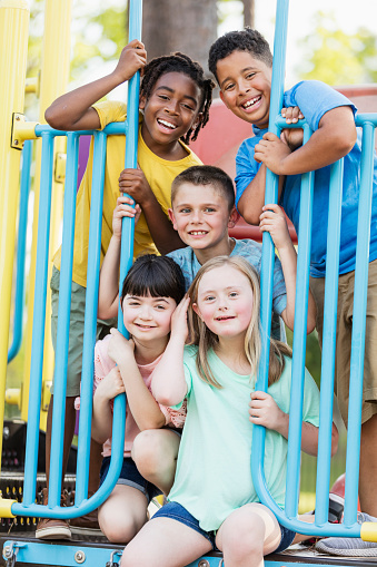 A multi-ethnic group of five children, 8 to 10 years old, playing together on a playground. They have climbed up onto the play equipment and are smiling at the camera. The girl with light brown hair, at the bottom, has down syndrome.