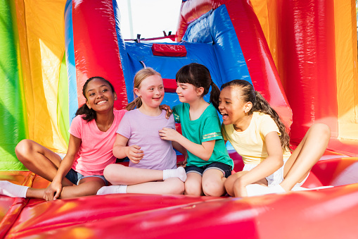 A multi-ethnic group of six girls sitting together in a bounce house, side by side, smiling and laughing. The girl second from the left has down syndrome.