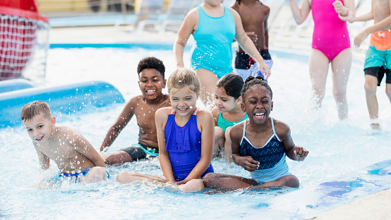 A multi-ethnic group of children, 7 to 10 years old, having fun together at a water park. Five of them are sitting in the water together, laughing and getting splashed.