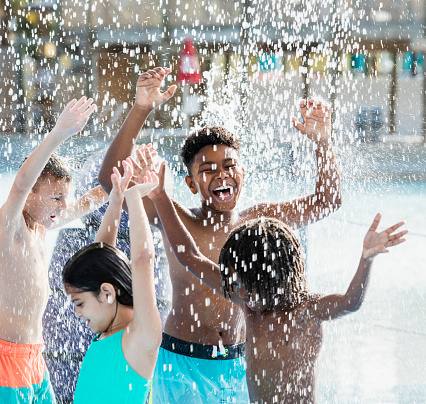 A multi-ethnic group of four children having fun at a water park, arms raised as they play under falling water on a sunny day. They are 8 to 10 years old.