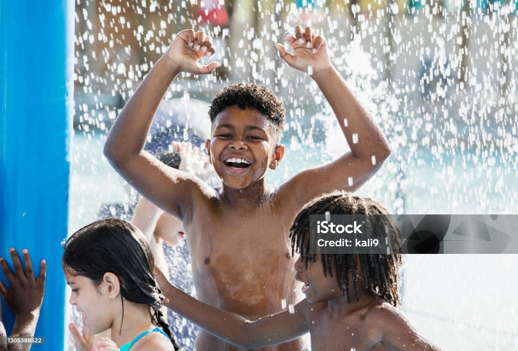 Boy with friends at water park A 10 year old African-American boy having fun playing at a water park. He is standing under splashing water with a group of friends, arms raised, laughing and looking at the camera. Water Park Stock Photo