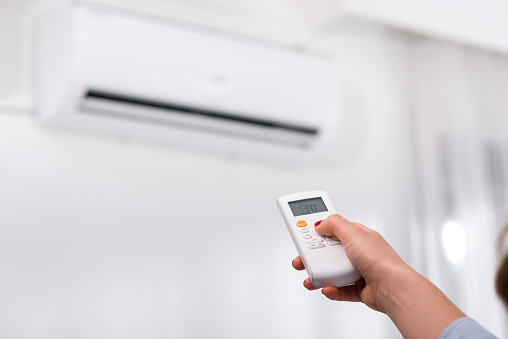 A young woman holding remote control, adjusting temperature of air conditioner mounted on a white wall. Indooor comfort temperature.