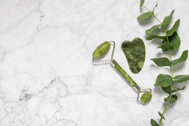 Green jade roller and gua sha stone for facial massage and eucalyptus branch on marble background. Home beauty and selfcare accessories. Face roller for anti age wrinkle treatment. Top view, flat lay. rodillo de jade verde y piedra gua sha para masaje facial y rama de eucalipto sobre fondo de mármol. accesorios para la belleza del hogar y el autocuidado, rodillo facial para el tratamiento antiedad de arrugas. vista superior, tendido plano.