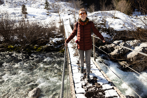Smiling woman crossing wooden bridge over a river in winter nature of Slovenia.