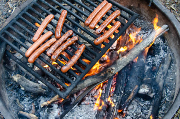 Roasting hotdog over open flame campfire. Grilling delicious meat hot dog over fire pit. Camping and cooking picnic lunch or dinner outdoors.