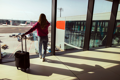 Back shot of a young woman standing next to an airport window while holding her suitcase