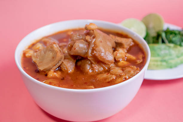 Delicious traditional Mexican Menudo on a white plate accompanied by a seasoned dish on a pink background.