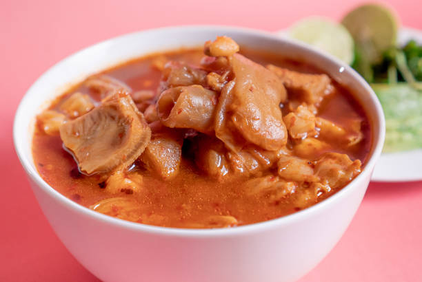 Delicious traditional Mexican Menudo on a white plate accompanied by a seasoned dish on a pink background.