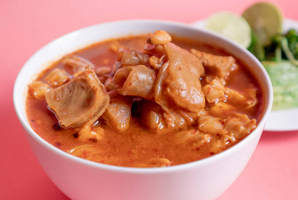 Delicious traditional Mexican Menudo on a white plate accompanied by a seasoned dish on a pink background.
