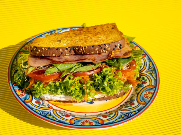 istock Exquisite ham sandwich with whole wheat bread, tomato, lettuce, avocado and cheese inside on a plate on a yellow table. 1305411751