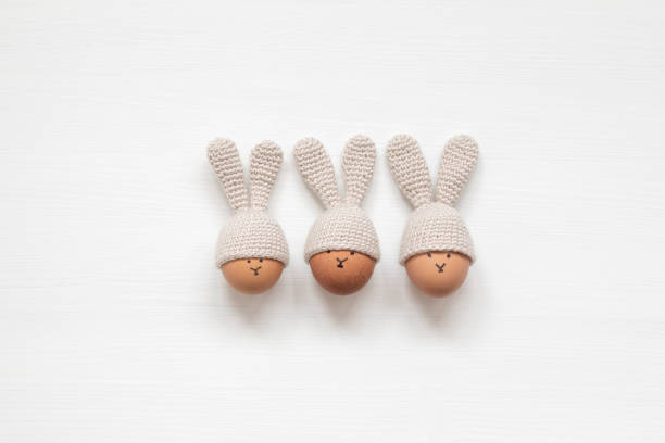 istock Three Easter eggs in crochet hats with bunny ears on white wooden background. Flat lay 1305436713