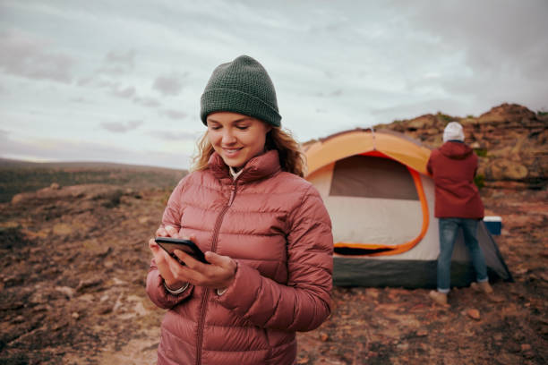 istock Happy young woman in winter clothing using smartphone during camping in mountain hill while man building tent in background 1305437243