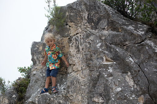 Child, blond toddler boy, standing on a big rock in the mountains, smiling happily