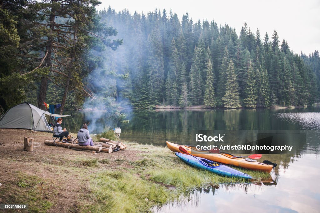Two female friends sitting next to campfire in a wild camp in the mountain. A shot of two beautiful woman having good time in a wild camping near to lake. Camping Stock Photo