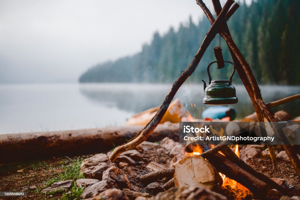 Shot of a cute vintage teapot in a campsite near to lake. Campfire with a vintage kettle next to the beautiful lake. Camping Stock Photo