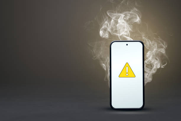 Smoking smartphone with failure icon A standing smartphone with smoke behind it and a warning symbol on the screen. Neutral background with copy space. overheated stock pictures, royalty-free photos & images