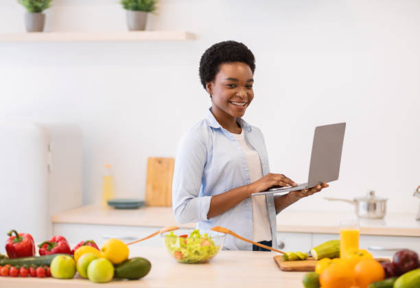 African Lady Holding Laptop Browsing Recipes Cooking Dinner In Kitchen