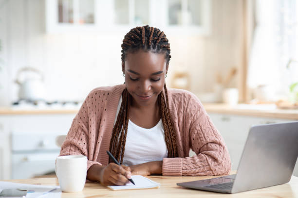 istock Smiling Black Student Woman Study With Laptop In Kitchen And Taking Notes 1305454119