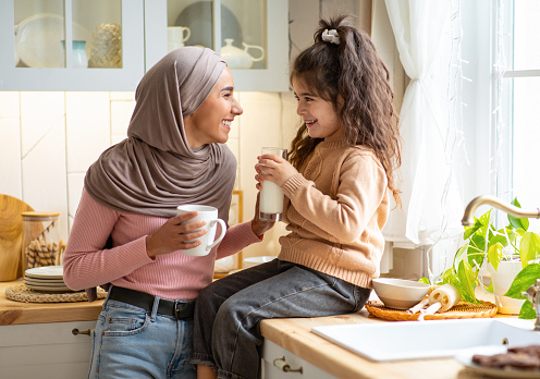 Happy Muslim Mom In Hijab And Her Little Daughter Bonding Together In Kitchen, Islamic Family Talking And Laughing, Mother Enjoying Coffee While Her Female Child Sitting At Table And Drinking Milk