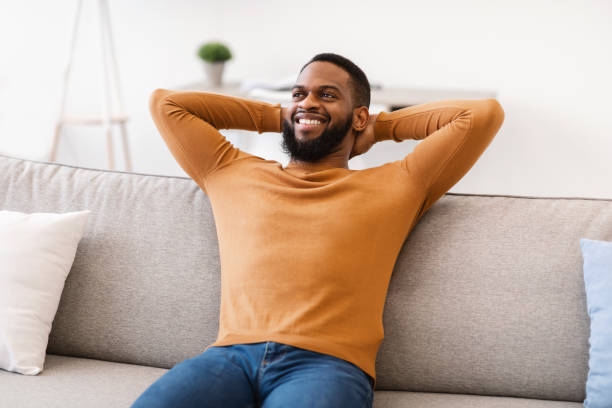 istock Black Guy Sitting On Couch Holding Hands Behind Head Indoor 1305455325