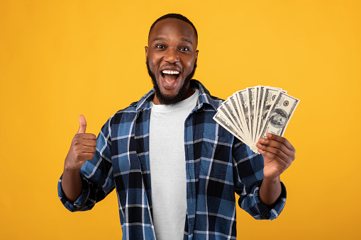 Big Luck. Joyful Black Guy Holding Money Cash Gesturing Thumbs Up Posing Standing Over Yellow Studio Background. Profit And Financial Success, Rich African Man Showing Like Gesture