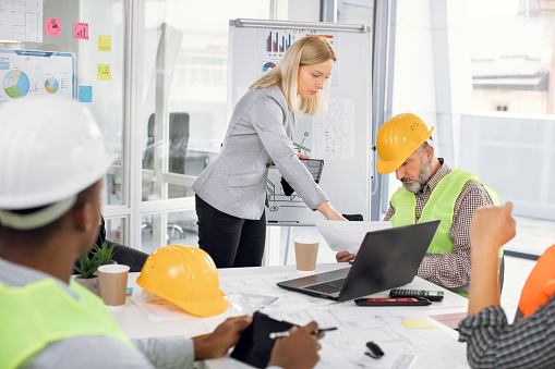 Group of coworkers in suits and helmets having business meeting at office. Responsible female engineer showing project of new building to mature male architect. Construction industry.