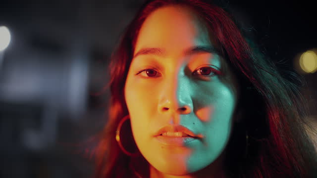 A portrait of a young woman lit by neon colored lights in the city at night.