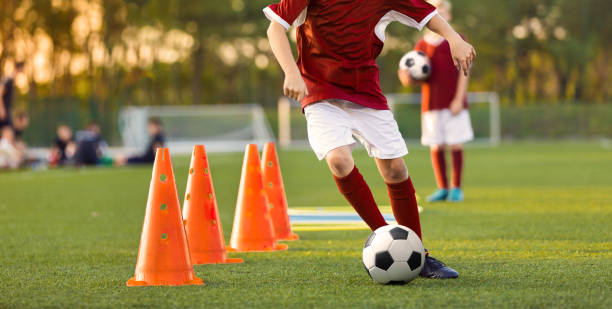 gruppe junger fußballer, die dribblings zwischen trainingskegeln üben. jungen kicken fußballbälle auf outdoor-training. jugendfußballmannschaft in roten trikots - mannschaftsfußball stock-fotos und bilder