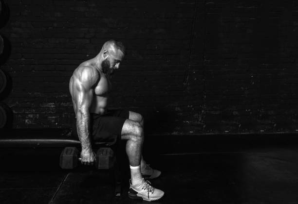 istock Young active strong sweaty fit muscular man with big muscles sitting on the bench and doing biceps workout with dumbbells in the gym as hardcore cross training 1305472507