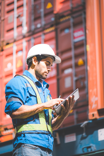 Engineer or foreman holding tablet and wears PPE to checking inventory or job details in the tablet with cargo container background. Engineering site and working with technology concept.