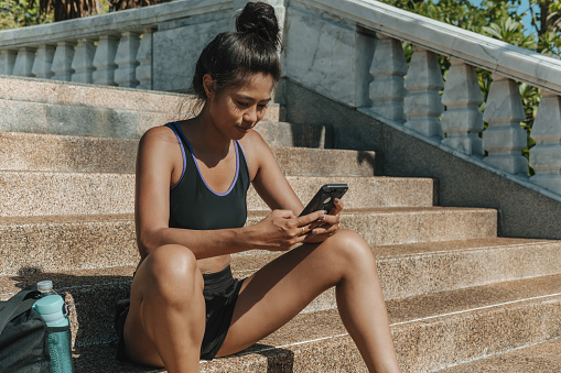 Sporty asian girl using smartphone for checking progress after training outdoors, typing message or calling friend online
