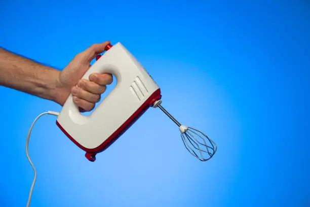 Electric food mixer held in hand by Caucasian male hand studio shot isolated on blue background.