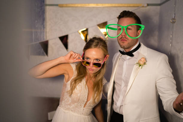 newlywed couple having fun in front of phto booth on her wedding reception party evening newly married wedding couple, bride in beautiful wedding dress, bridegroom in white dinner jacket, enjoying party and taking pictures at photo booth in the evening photo booth stock pictures, royalty-free photos & images