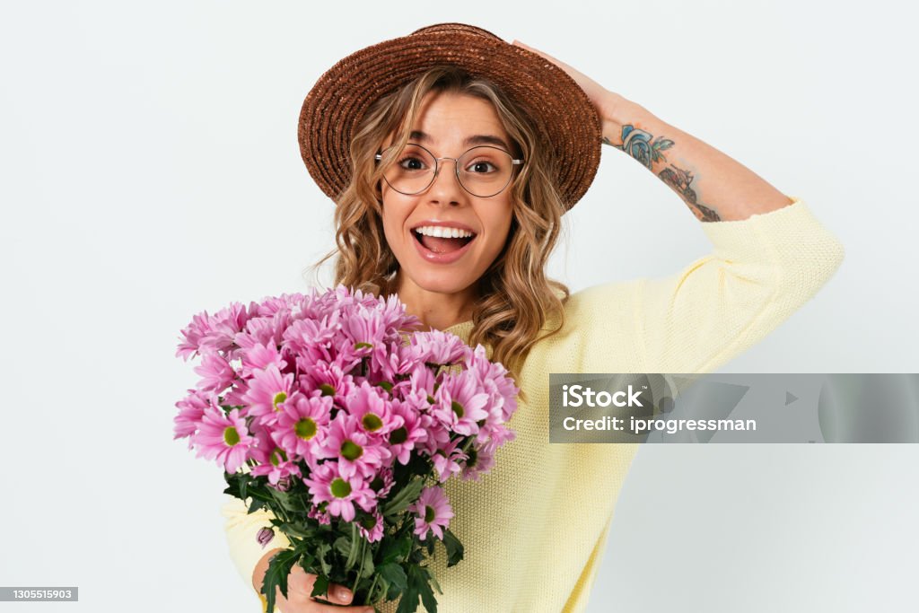 Cheerful young woman in summer straw hat Cheerful young woman in summer straw hat and eyeglasses holding bouquet of pink flowers standing on white background. Springtime Stock Photo