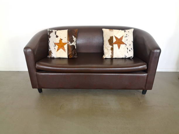 Leather sofa, brown, with two decorative pillows on a beige-brown floor in front of a white wall.