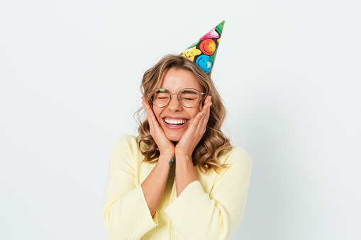 Joyful emotions, young woman in birthday cap closed her eyes with happiness holding her face smiling while standing on white studio background.