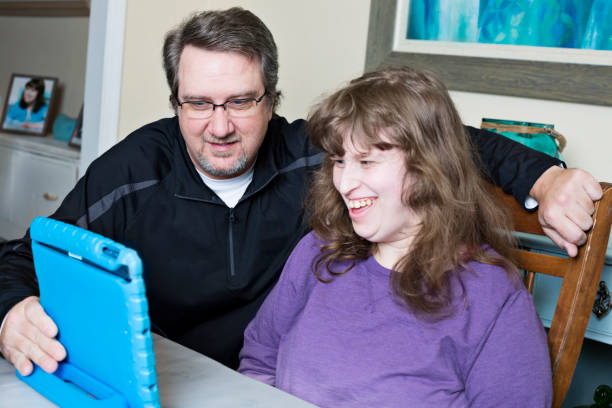 Father assists disabled daughter with her online homework via the digital tablet Dad has to assist his intellectually disabled daughter during virtual school. They both smile at the digital tablet as the teacher communicates to both of them what today's lesson plan will be. special education stock pictures, royalty-free photos & images