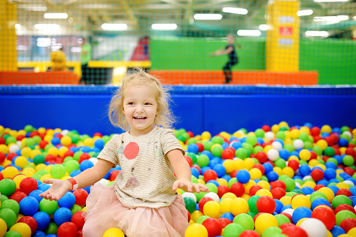 Curly little girl having fun in ball pit with colorful balls. Child playing on indoor playground. Kid jumping in ball pool.