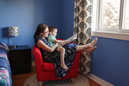 Mother with son boy sitting in armchair at home and reading book together. Child kid early development education. Family mom and baby spending time together. Homeschooling concept.
