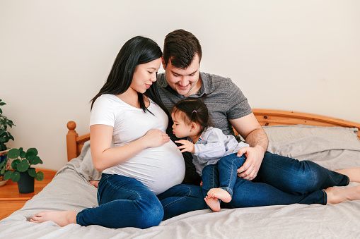 Family Asian Chinese pregnant woman and Caucasian man with toddler girl sitting on bed at home. Mother, father and baby daughter expecting waiting for new family member. Ethnic diversity.