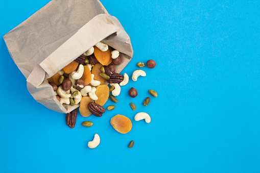 Mixture of different nuts and dried apricots in a paper bag  on the blue background. Healthy snack. To view. Copy space.