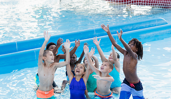 A multi-ethnic group of children, 7 to 10 years old, having fun together at a water park, standing together in a pool, reaching up to try to catch a ball.